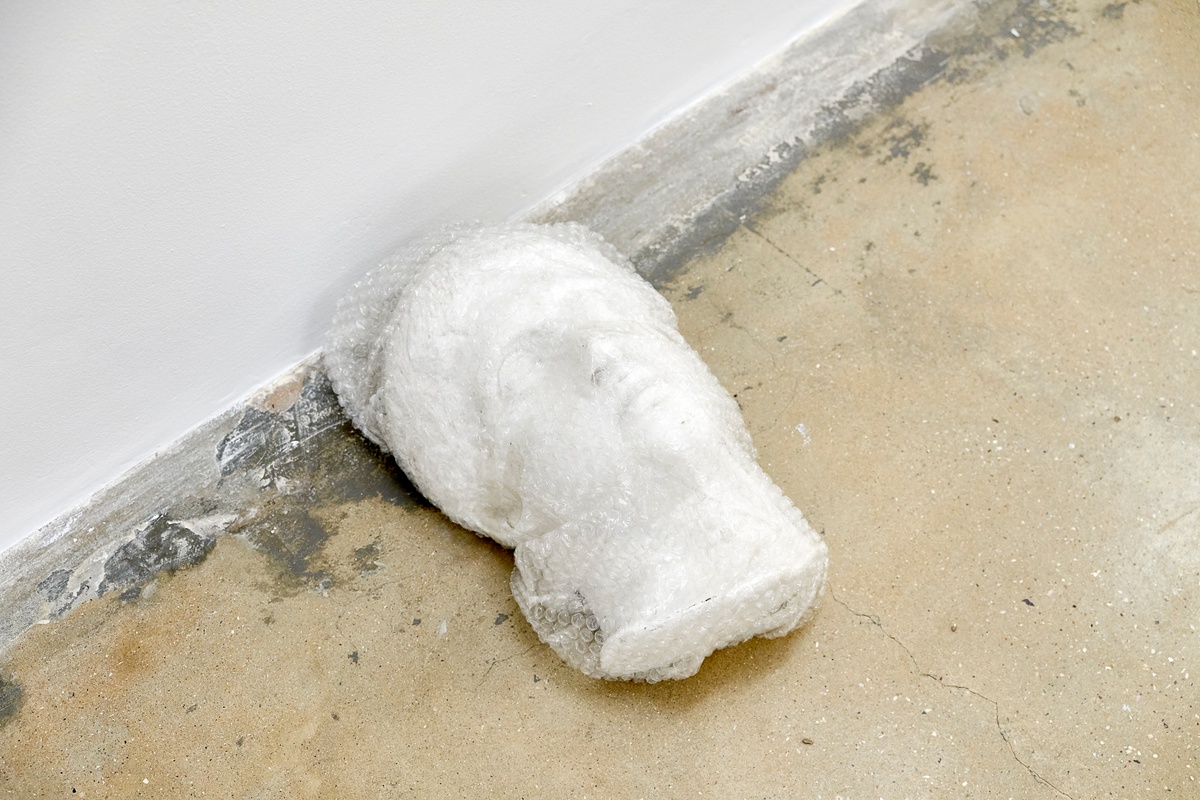 Installation photograph from the 2018 rendition of ‘Parallel Play’ in A4’s Gallery. In the middle, a bust wrapped in bubble wrap from Rodan Kane Hart’s ‘Western Death Mask(s)’ project lays at the base of a wall.
