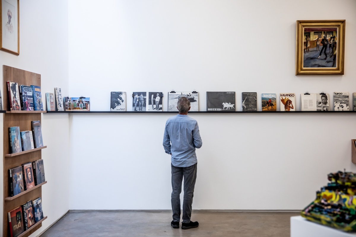 Event photograph from the opening of the Photo Book! Photo-Book! Photobook! exhibition in A4’s Gallery in an area dedicated to photobooks from the years 1967 to 1994. In the middle, an attendee looks at a display shelf. On the right, Gerard Sokoto’s oil painting ‘Street Musician in District Six’ hangs above the shelf.
