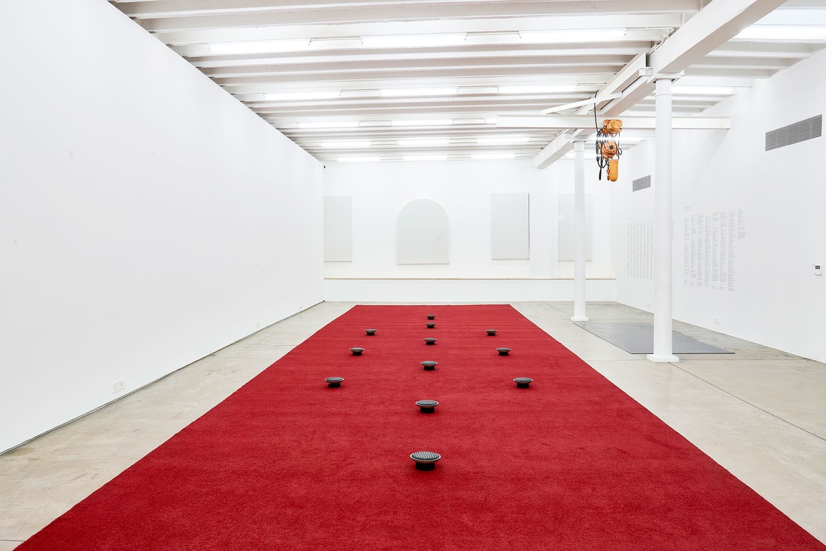 Installation photograph from The Future Is Behind Us exhibition in A4’s Gallery. In the middle, James Webb’s sound installation ‘Prayer’ features an long red carpet with upturned audio speakers laid out in a geometric pattern.
