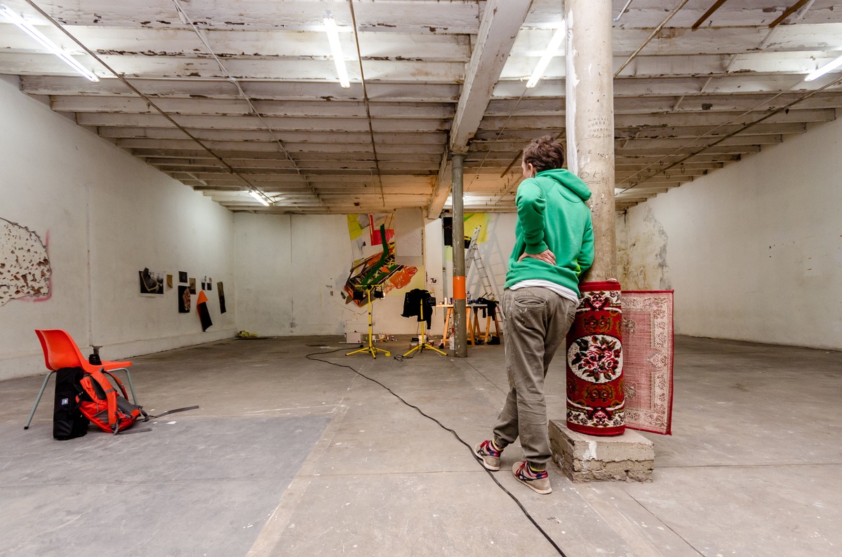 Process photograph from Dorothee Kreutzfeldt’s residency on A4’s 1st floor. At the front, Kreutzfeldt leans against a concrete support beam. At the back, the wall features a painted mural.
