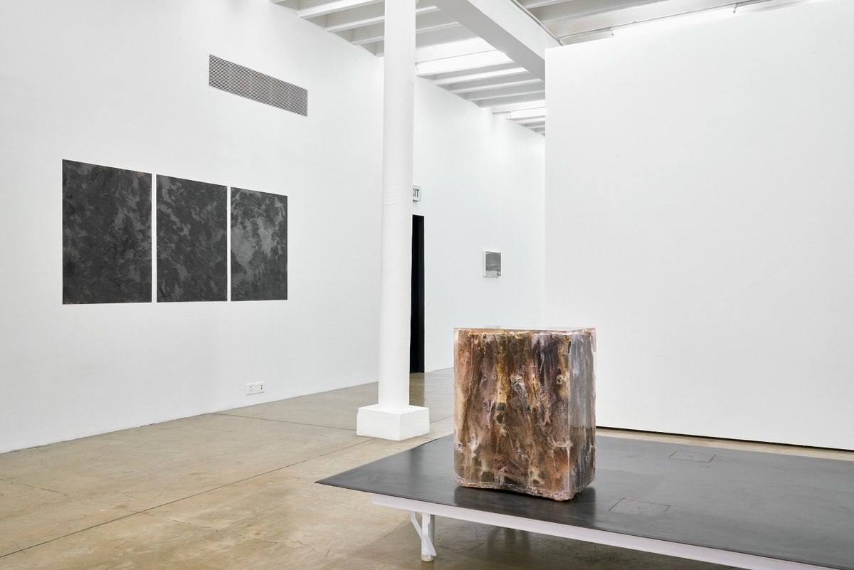 Installation photograph from the ‘without a clear discernible image’ exhibition in A4’s Gallery. On the left, three of Kevin Beasley’s graphite, ink and acrylic medium works are mounted on the gallery wall. On the right, Beasley’s untitled ebony and resin sculpture sits on a metal platform.
