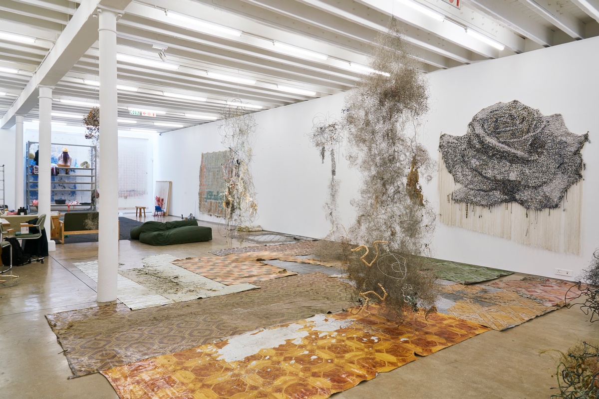 Process photograph from ‘Open Production’, Igshaan Adams’ hybrid studio/exhibition in A4’s Gallery. At the front, sheets of found nylon flooring with various patterns line the gallery floor, with one of Adams’ wire mixed media sculptures suspended from the ceiling above it. On the right, Adams’ mixed media tapestry ‘Al Mumeet (The Bringer of Death/The Destroyer)’ is mounted on the wall. 
