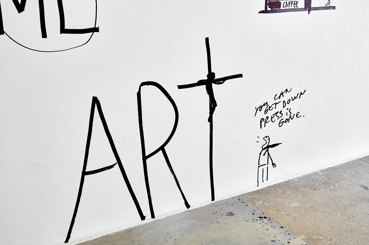 Installation photograph from Dan Perjovschi’s ‘The Black and White Cape Town Report’ exhibition in A4’s Gallery. On the left, the word ‘ART’ is written in black felt pen marker at the base of the wall, with the letter T drawn to resemble a crucifix. On the right, a small humanoid figure points away from the word with the text “you can get down, press is gone ..” written above its head.
