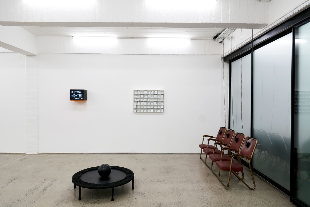 Installation photograph from the ‘Model’ exhibition in A4’s Reading Room. At the front, Bogosi Sekhukhuni’s ‘Dark Gravity,’ consists of a black trampoline with a black bowling ball resting on it. At the back on the wall, DB Amorim’s video ‘remoting’ is on the left, and Kyle Morland’s paper and tape sculpture ‘Library of Forms’ is on the right. On the right, Bhavisha Panchia’s sound work ‘Imagine you’re in a museum: What do you hear?’ features four chairs with headphones.

