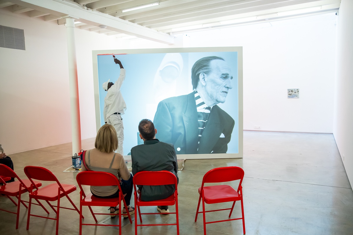 Event photograph from the preview of the 'A Little After This' exhibition in A4 Arts Foundation that shows Alex Da Corte's video installation 'ROY G BIV'. At the back, a large white wooden box with a back-projected screen plays Da Corte's video as an individual dressed in white overalls repaints the box red. At the front, 7 red powder-coated viewing chairs are arranged in an arch, with attendees seated there.
