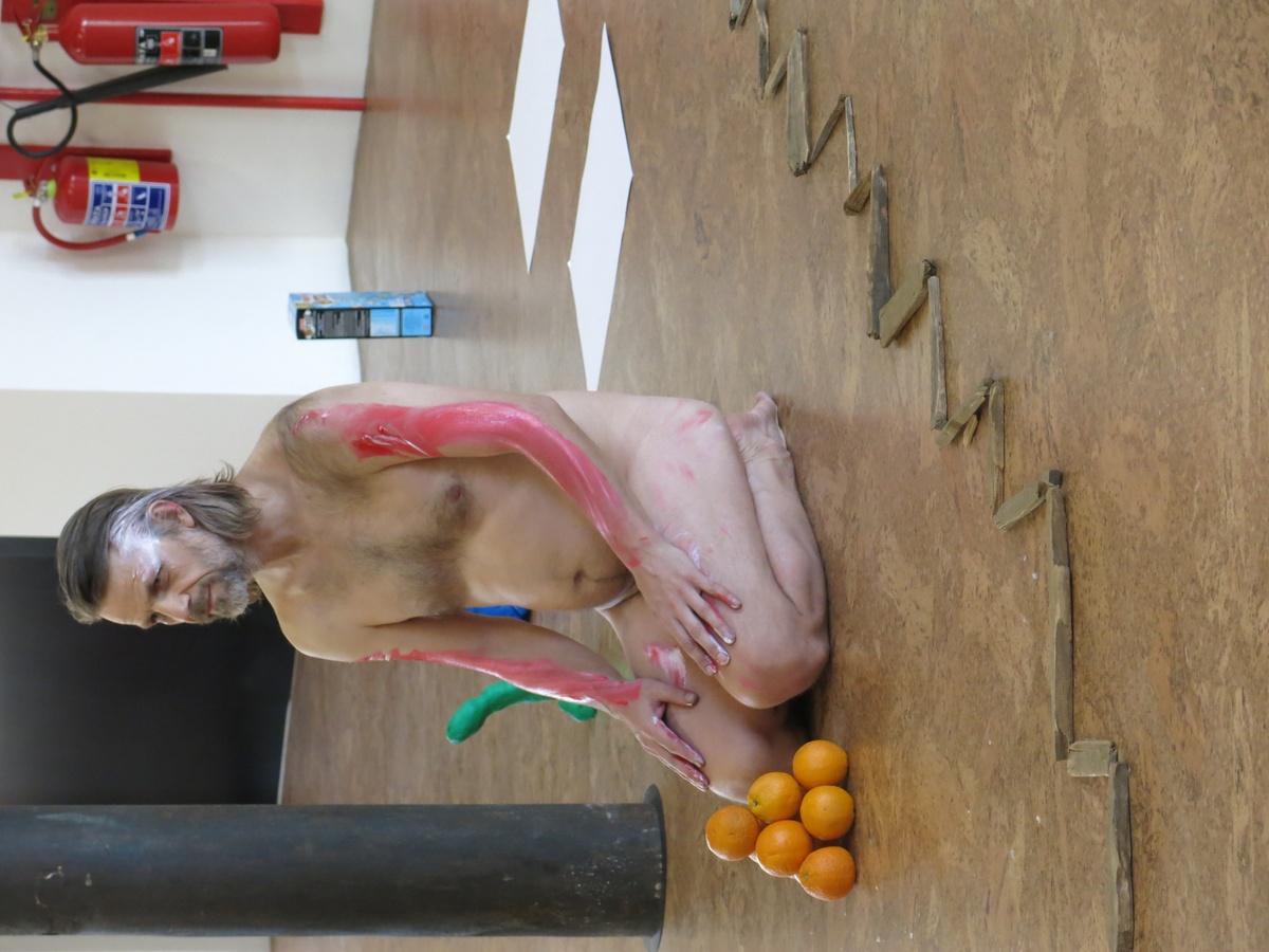 Event photograph from Mårten Spångberg’s solo dance ‘Digital Technology’ on A4’s top floor. In the middle, Spångberg is seated unclothed in a kneeling position on the floor, with paint on the side of his face and along the top of his arms. At the front, a pyramid of oranges sits on the floor.
