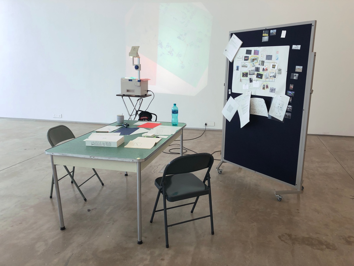 Process photograph from the More For Less exhibition in A4’s Gallery that depicts a makeshift curatorial studio that consists of a table, a pinboard and an overhead projector.
