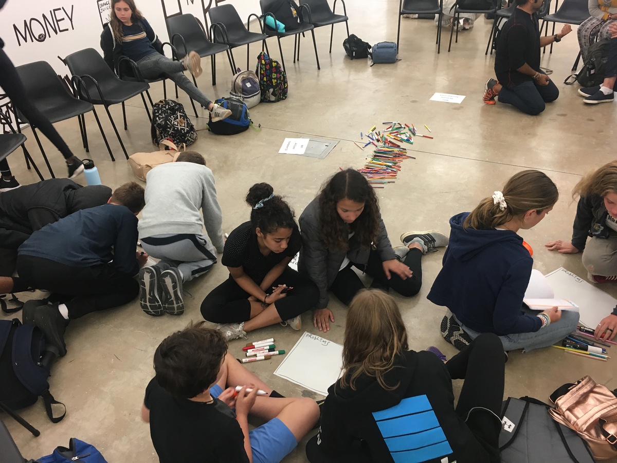 Photograph from the ‘Water Workshop with Edu Africa’ exchange in A4’s Gallery. At the back, rows of chairs surround an open area with variously coloured felt pen markers laying on the floor. At the front, students are huddled in groups around pages on the gallery floor.
