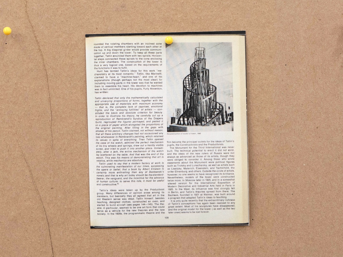 Installation photograph from Mitchell Gilbert Messina's residency in A4 Art Foundation shows a wall-mounted strip of cardboard hosting pinned research notes. A 1968 reconstruction of Vladimir Tatlin’s _Monument for the Third International_, the original of which was destroyed in 1920. The page is taken from MOMA’s catalogue for _The machine, as seen at the end of the mechanical age_.
