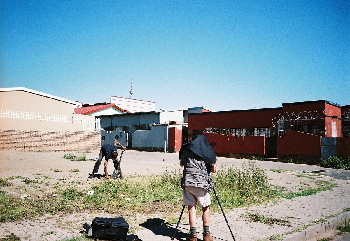 Still frame from the documentary on David Goldblatt directed by Daniel Zimbler. On the left, an individual with a spade removes weeds from a pavement. On the right, Goldblatt operates a camera on a tripod.
