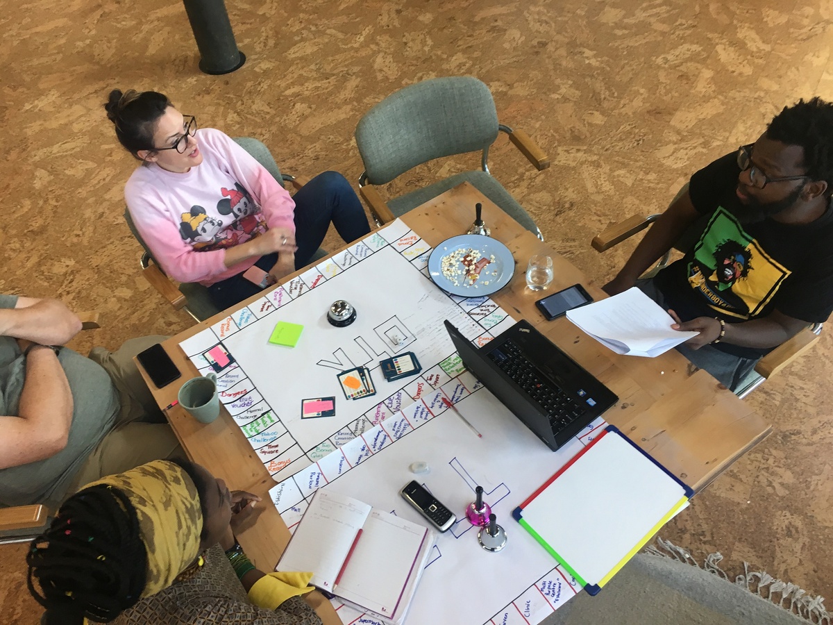 Event photograph from part 2 of Jefferson Bobs Tshabalala’s residency on A4’s top floor. A topdown view shows a wooden table that hosts game boards drawn out with felt pen markers on sheets of paper. Four participants sit in discussion around the table.
