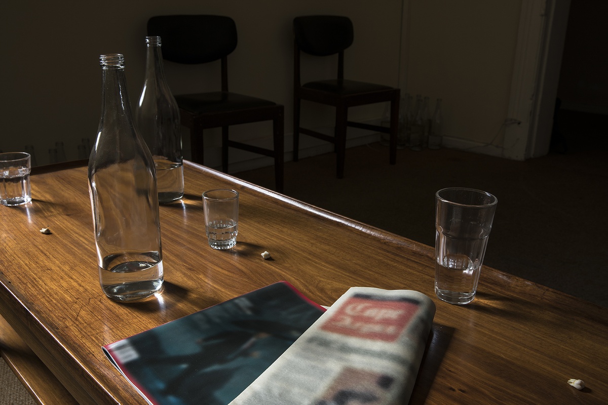 Installation photograph from Gain Maria Tosatti’s offsite exhibition ‘My Hart is so Leeg soon ’n Spieël (My Heart is a Void, the Void is a Mirror)’ that depicts the top of a wooden coffee table. Towards the back, drinking glasses and glass bottles with small amounts of water in them, along with human teeth. Towards the front, reproductions of a magazine and a newspaper, with image and text blurred so as to be illegible. 
