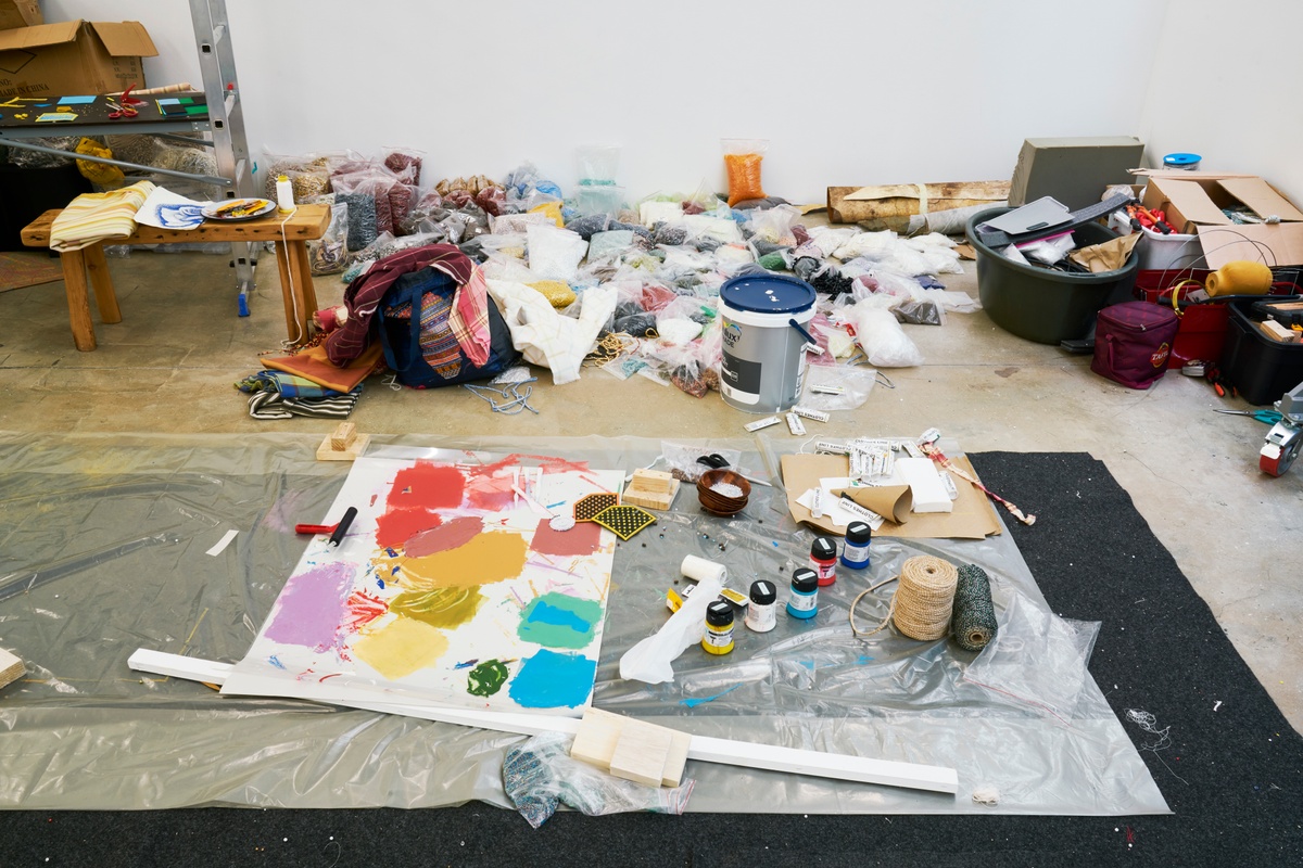 Process photograph from ‘Open Production’, Igshaan Adams’ hybrid studio/exhibition in A4’s Gallery. At the front, plastic sheeting lines the gallery floor, covered in paper for testing colours, jars of dye and rolls of nylon rope. At the back, a large pile of variously coloured and sized beads in resealable plastic bags.
