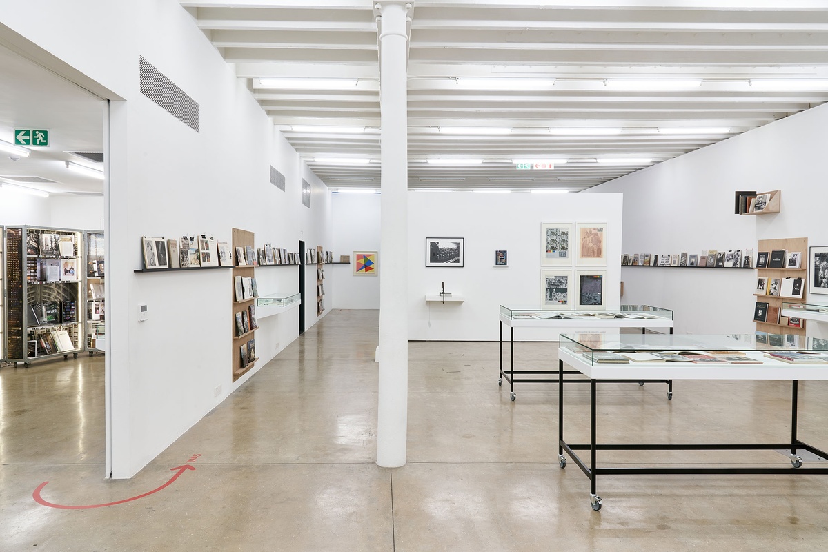 Installation photograph from the Photo Book! Photo-Book! Photobook! exhibition in A4’s Gallery. At the front, two freestanding display cases with printed matter. In the middle, a moveable gallery wall that features Peter Magubane’s photograph ‘Mine recruits humiliated by being forced to strip naked for tuberculosis examination, Witwatersrand (1968)’, Jacobsens Publishers’ book ‘Index of Objectionable Literature’, Ernest Cole’s book ‘House of Bondage’ and four screenprints from Gavin Jantjes’ ‘A South African Colouring Book’ series. On the left, shelving units with printed matter from the years 1945 to 1967 line a white wall, with Albert Newall’s oil painting ‘Harmonic Development within a Square’ mounted on a wall at the back. On the right, shelving units with printed matter from the years 1967 to 1994 line a white wall, along with David Goldblatt’s photograph ‘Climbing into kibbles at the shaft-head, the shift gets ready to go down. President Steyn No.4 shaft, Welkom, Orange Free State. 1969’.
