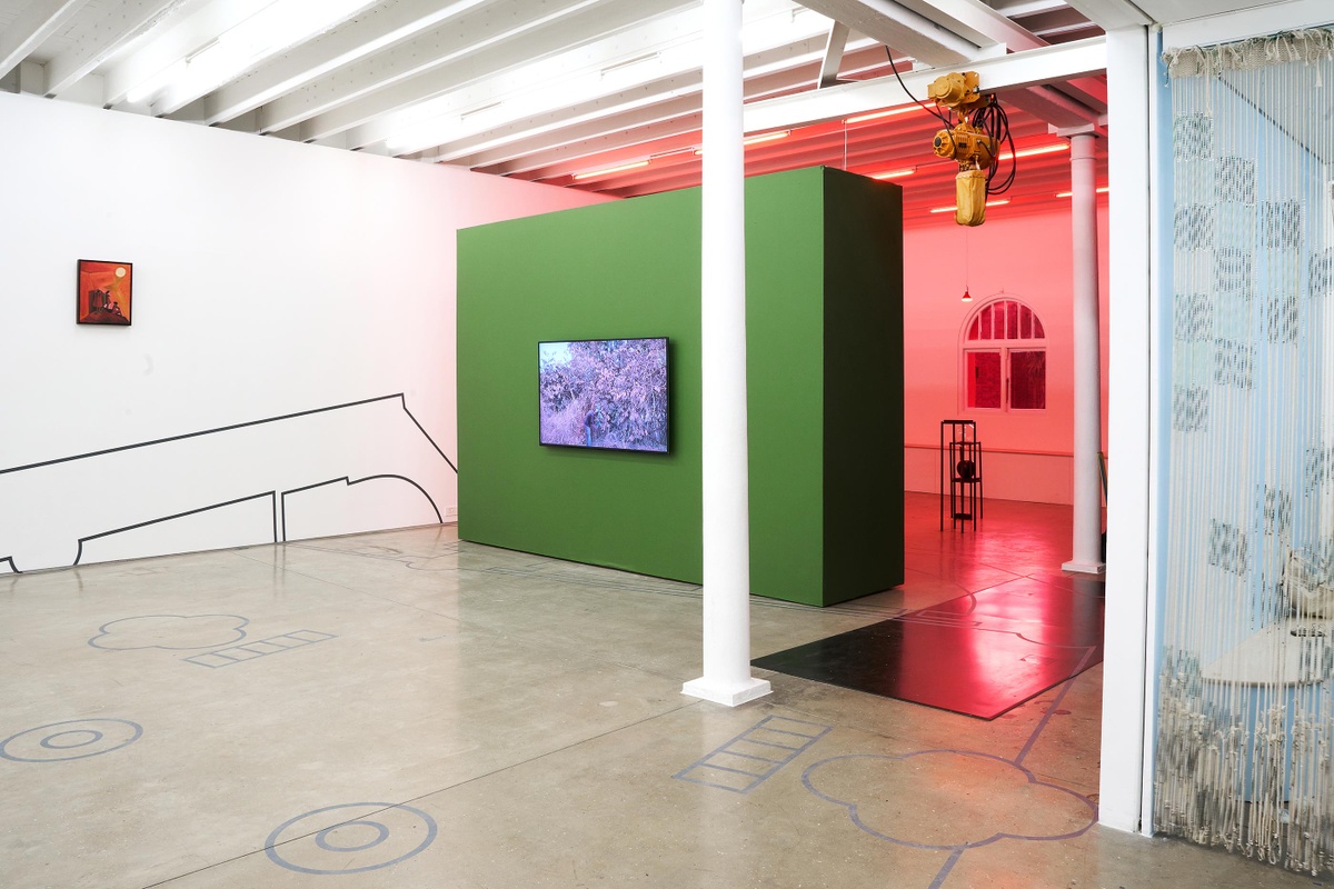 Installation photograph from the Customs exhibition in A4’s Gallery that shows Dor Guez’s vinyl installation ‘Double Stitch’ stretching across the gallery wall on the left and the gallery floor below. On the left, Peter Clarke’s acrylic painting ‘Anxiety’ is mounted on a white gallery wall. In the middle, Kapwani Kiwanga’s video work ‘Vumbi’ plays on a screen mounted on a green gallery wall. At the back, steel armatures from Nolan Oswald Dennis’ installation ‘garden for fanon’ sits on the gallery floor. On the right, Igshaan Adams’ tapestry ‘Al asma ul Husna’ is suspended from the ceiling.
