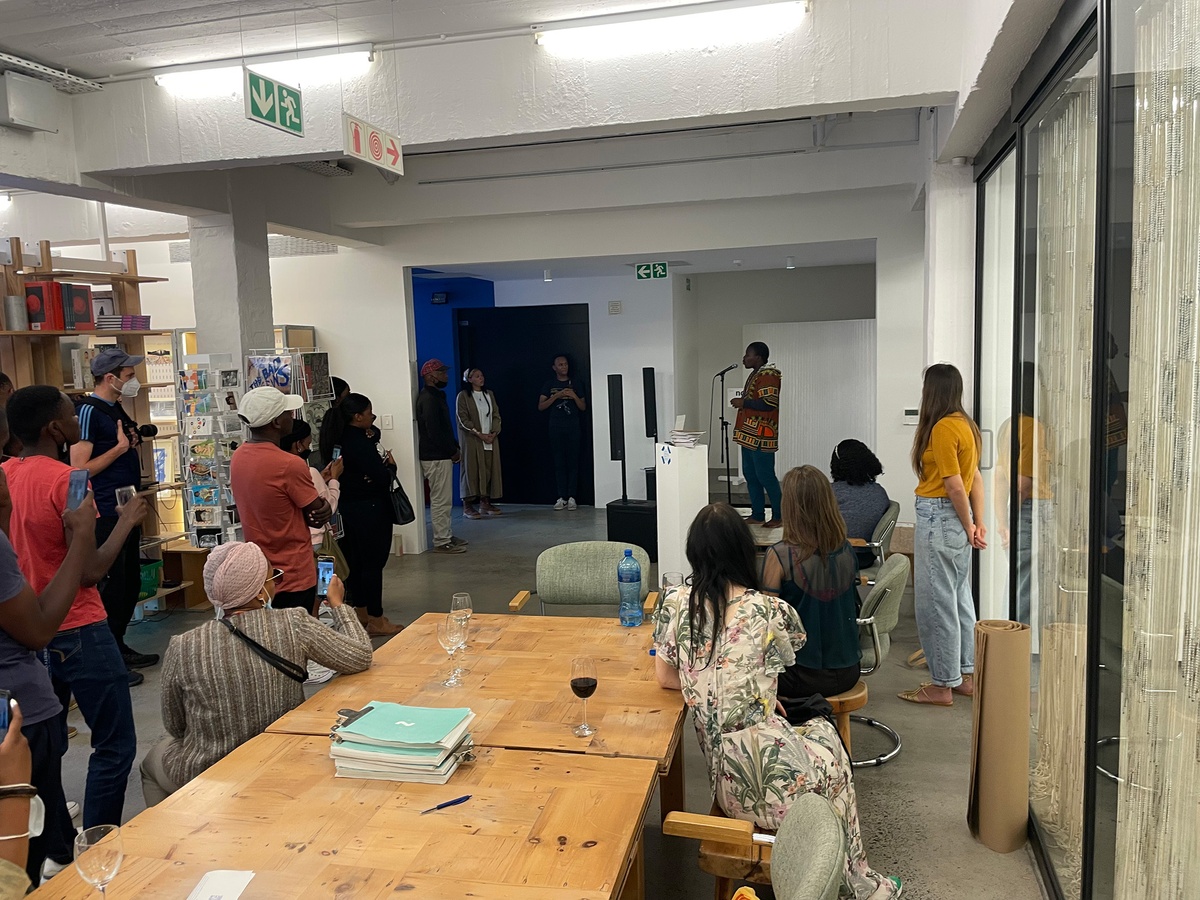 Event photograph from the ‘Umcimbi’ exchange in A4’s Goods project space. At the back, a individual performs into a mic. At the front, attendees sit at a table. On the left, attendees stand listening.
