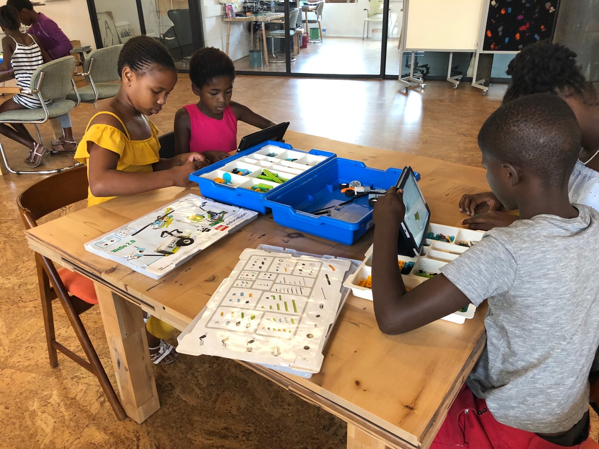 Event photograph from the Lego Robotics workshop by ORT SA CAPE on A4’s top floor. A table laden with lego electronic components in plastic containers is surrounded by children operating tablet computers.
