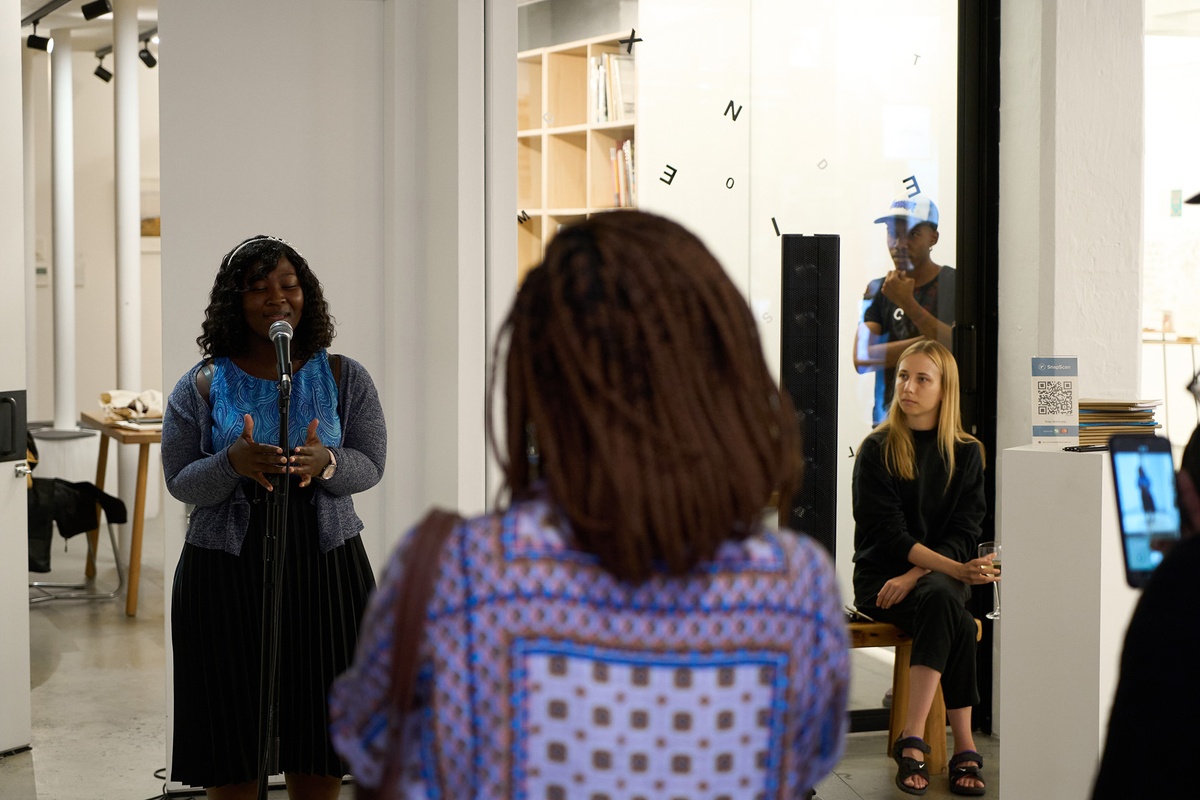 Event photograph from the ‘Umcimbi’ exchange in A4’s Goods project space. On the left, a participant performs into a mic. On the right, A4’s Nkhensani Mhkari and Lucienne Bestall watch the performance.
