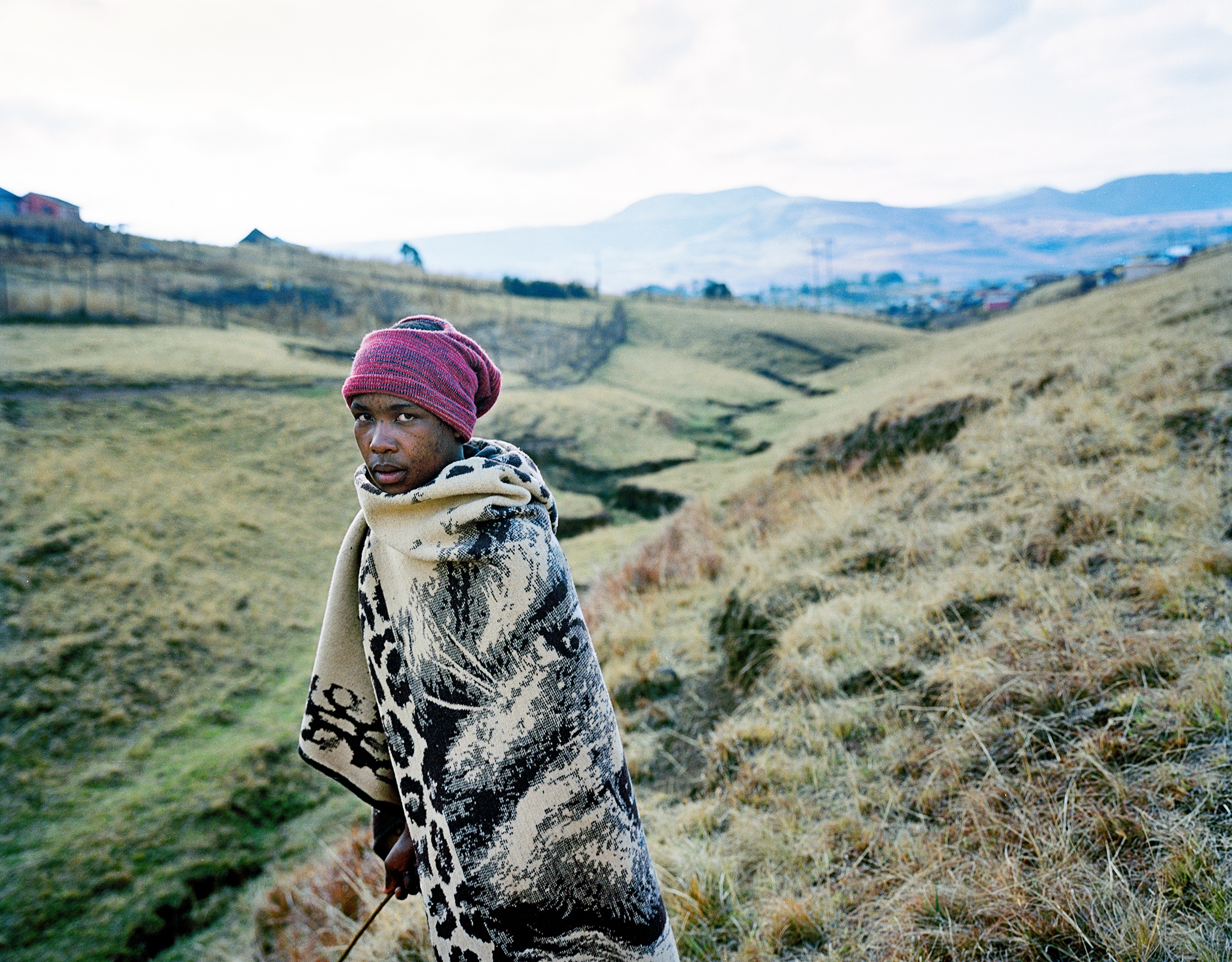 Lindokuhle Sobekwa's photograph 'Melusi wenkomo' shows an individual wrapped in a blanket standing in front of a grassy hillside.
