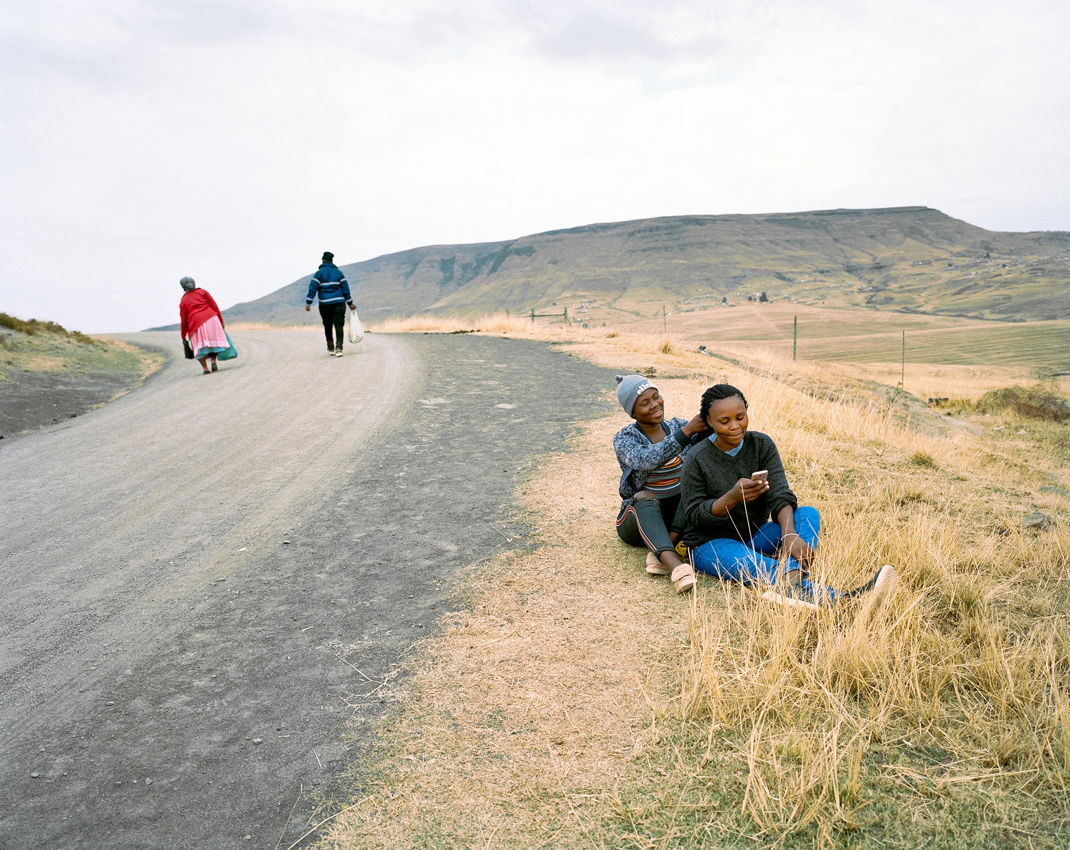 Lindokuhle Sobekwa's photograph 'Estopin Qumbu' shows two individuals walking on a dirt road on the left, and two individuals sitting on the grassy roadside on the right.
