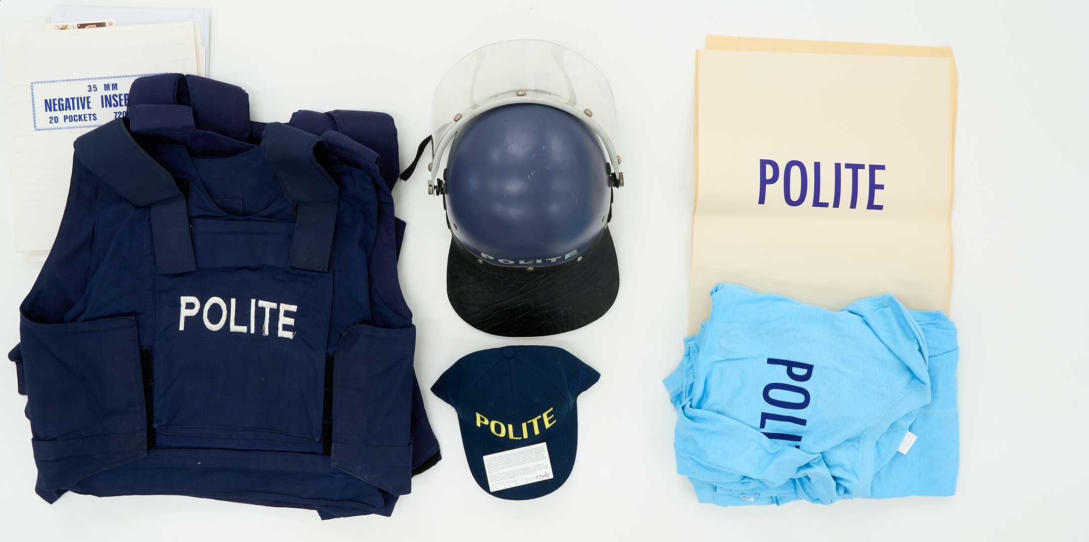 Photograph of paraphernalia from Christian Nerf’s ‘Polite Force,’ unpacked on the occasion of the 2022 Investec Cape Town Art Fair. The topdown view features a vest, helmet, cap, t-shirt and poster.
