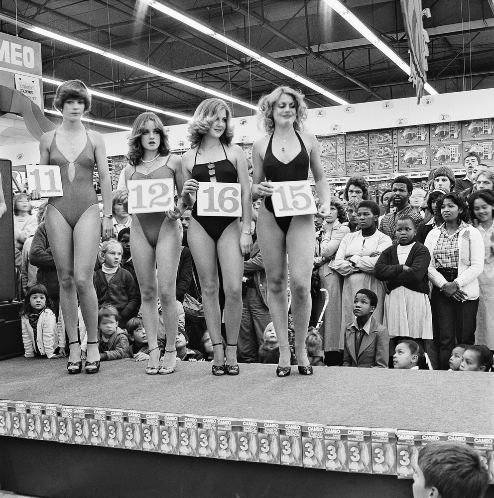 David Goldblatt's monochrome photograph 'Saturday morning at the Hypermarket: Semi-final of the Miss Lovely Legs Competition. 28 June 1980' shows a row of individuals standing on an indoor platform. The are wearing swimming clothes and placards printed with identification numbers.
