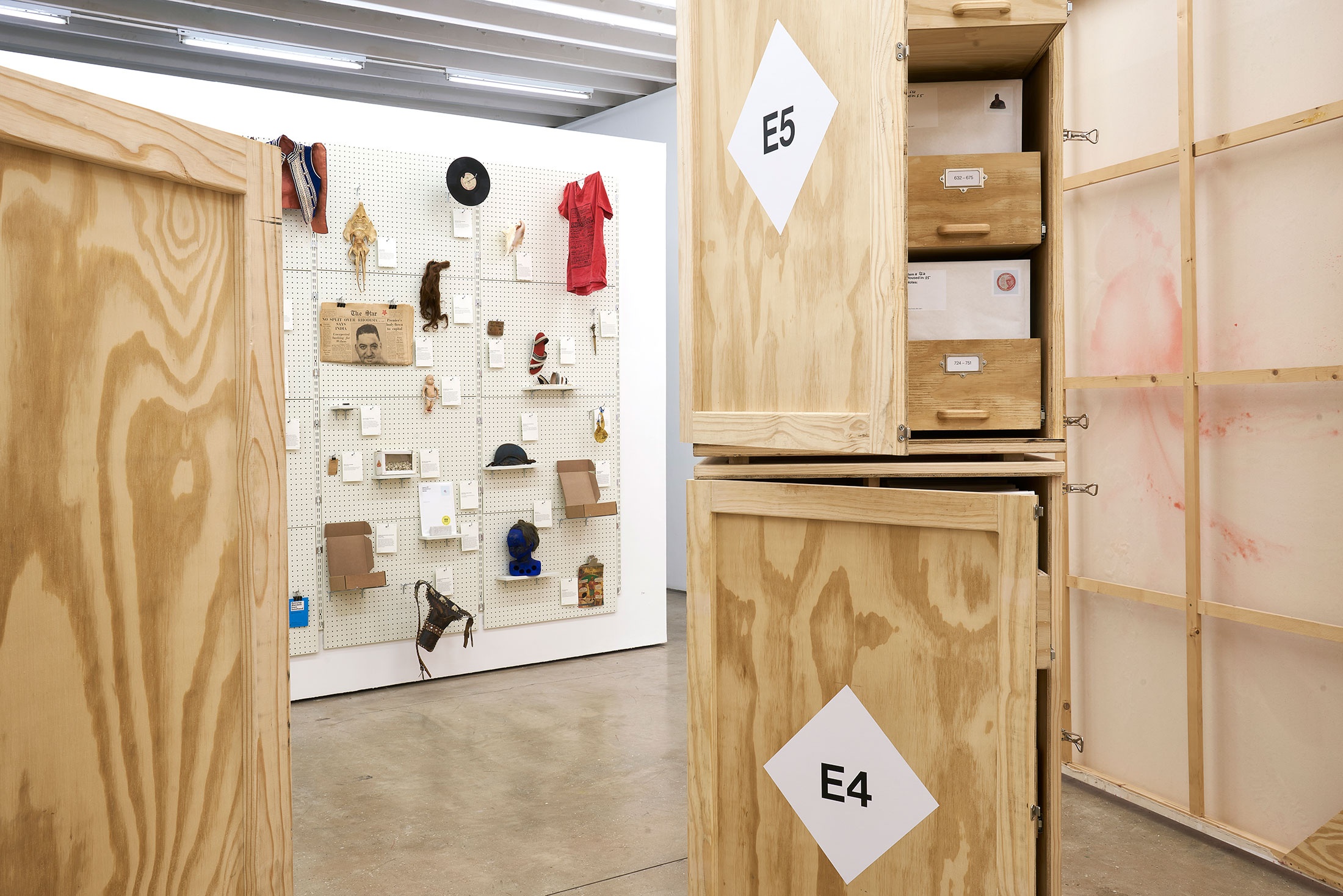 Installation photograph from the 'A Little After This' exhibition in A4 Arts Foundation's gallery. At the back, collected objects from Penny Siopis' 'Will' work mounted on a white moveable gallery wall through the use of perforated metal panels. At the front, collected objects from Penny Siopis' 'Will' work organised in envelopes, which in turn are arranged in bespoke wooden filing cabinets.
