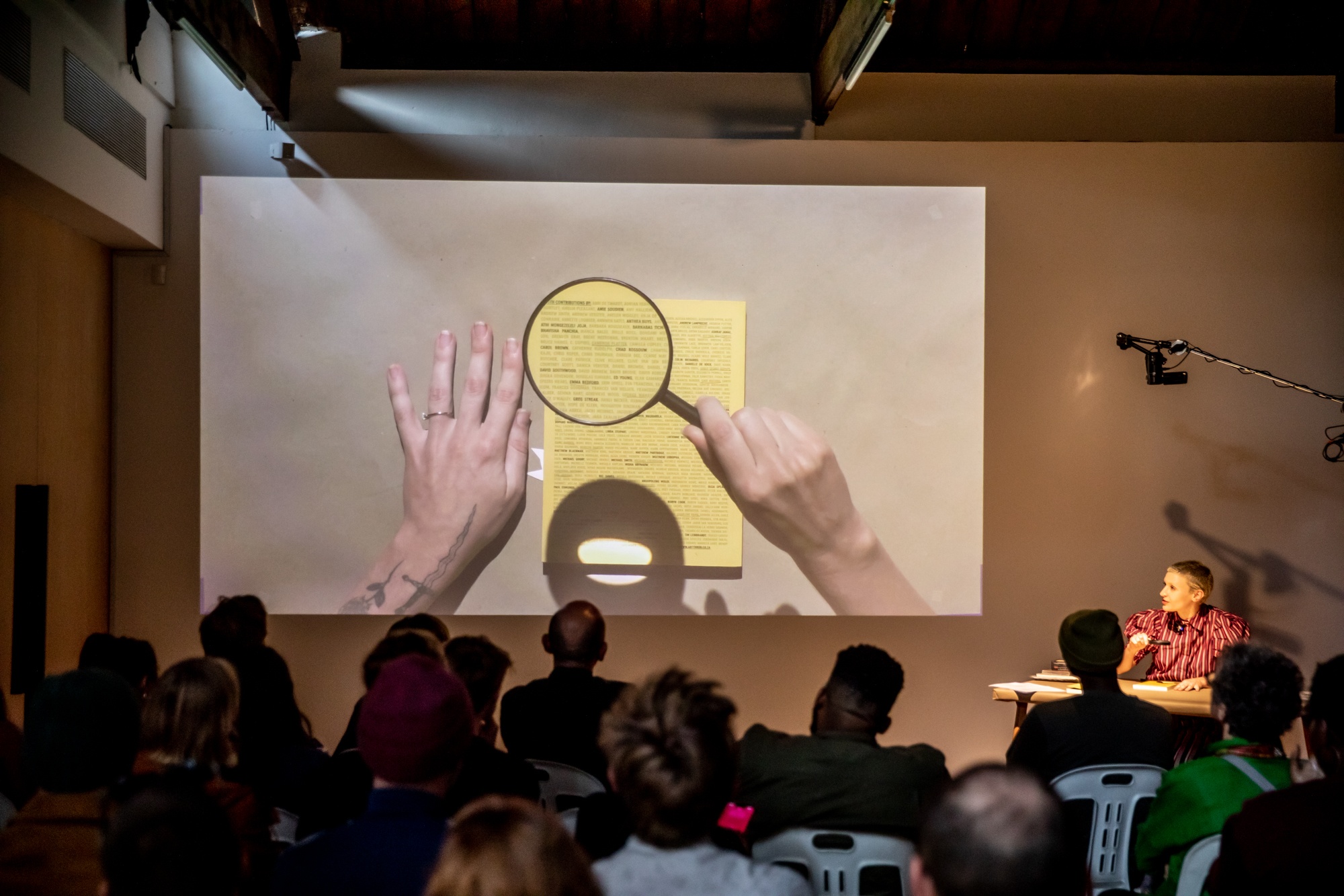 Event photograph from the book launch of 'ArtThrob: 25 Years of Art Writing in South Africa' in A4 Arts Foundation. On the right, writer Keely Shinners is seated at the speaker's table. On the left, a livestream top-down view of the speaker's table is projected onto the wall.
