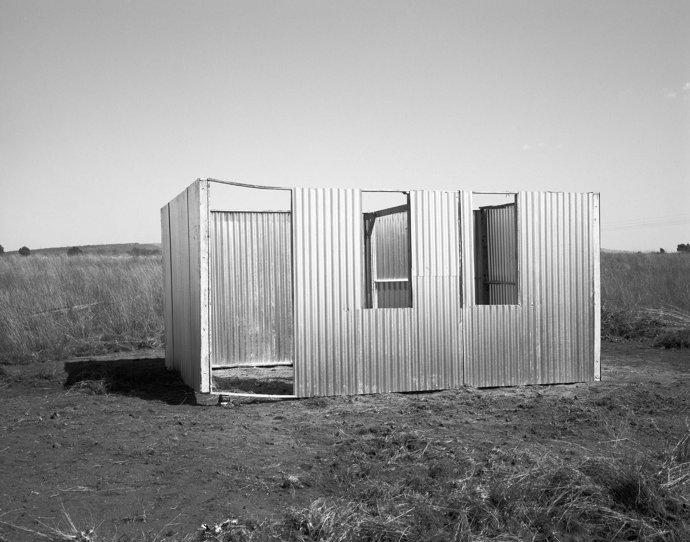 David Goldblatt's monochrome photograph's monochrome photograph 'A new shack under construction, Lenasia Extension 9, Johannesburg' shows a partial house structure composed of corrugated metal.
