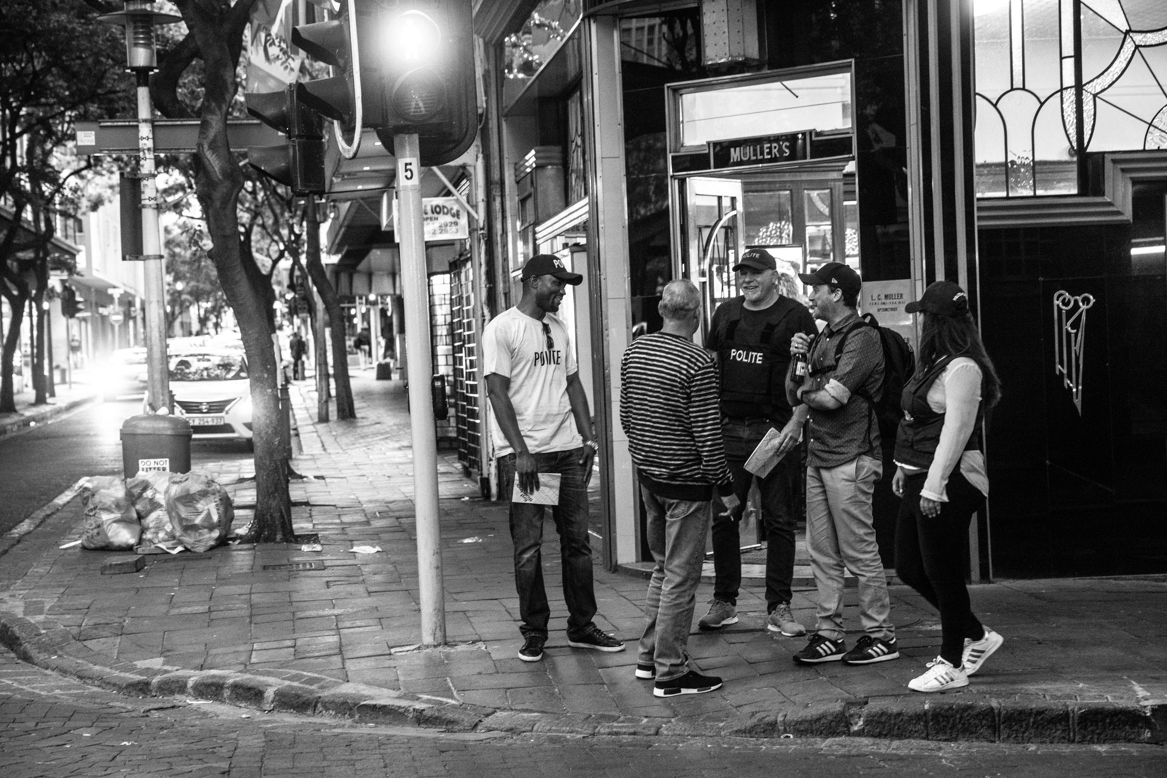 Monochrome process photograph from the 2019 rendition of Christian Nerf’s performance piece ‘Polite Force.’ On the right, two participants dressed in ‘Polite Force’ riot gear interact with member of the public on a street corner.
