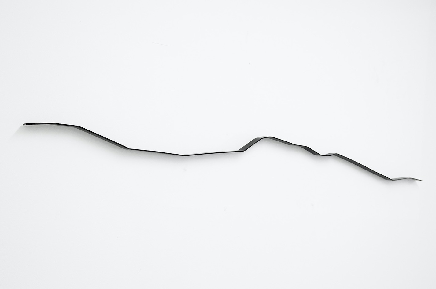 Installation photograph from the ‘Sounding the Void, Imaging the Orchestra V.1’ exhibition in A4’s Gallery. Kyle Morland's black passivated mild steel sculpture 'Ruler' is mounted on a white wall.
