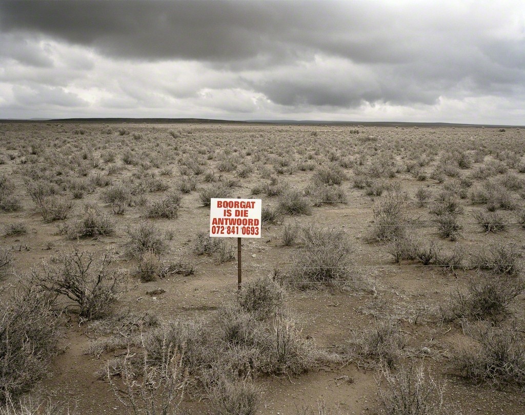 David Goldblatt's colour photograph 'Boorgat is die Antwoord, De Brak, on the Fraserburg-Sutherland road. Western Cape' shows a white sign with red text in a field of dry shrubs.
