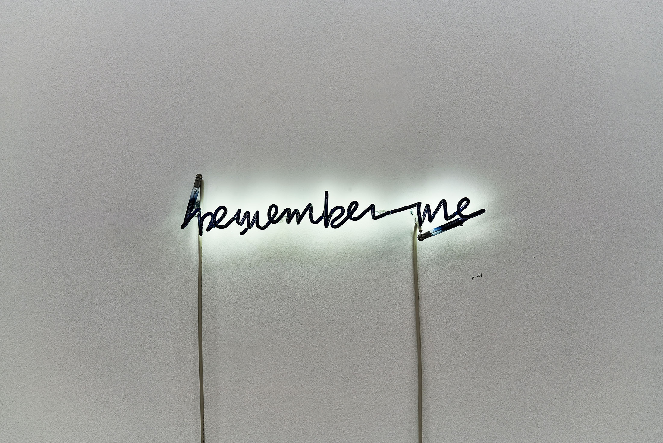 Installation photograph of Steve McQueen’s neon tube and acrylic paint sculpture ‘Remember Me,’ from the Customs exhibition in A4’s Gallery, that shows the phrase “remember me” described in neon tubing with the front covered over with acrylic paint and mounted on a white wall.

