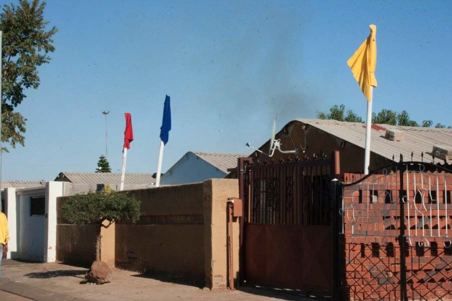 Process photograph from ‘Extracting Colour’, Mbali Dhlamini’s offsite residency. A brickwork building features three flagpoles, with flags in yellow, blue and red respectively.
