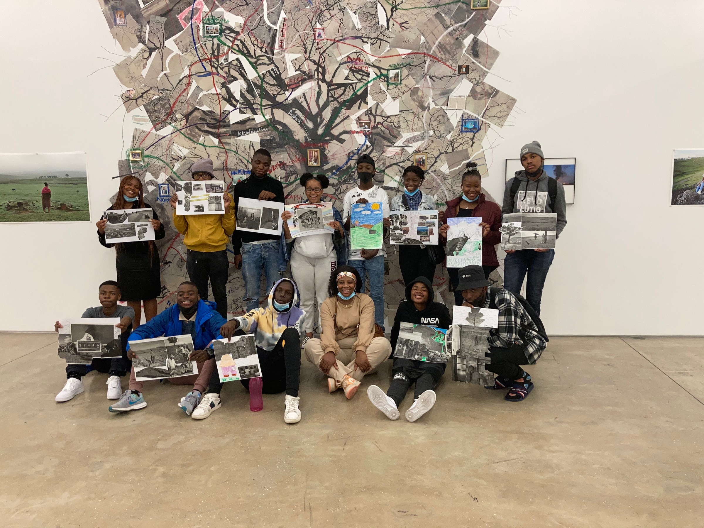 Event photograph from the ‘Lalela x A4' exchange during the ‘Tell It to the Mountains’ exhibition in A4’s Gallery. At the front, students from Lalela’s after-school programme stand in two rows with artworks they created in response to the exhibition. At the back, Lindokuhle Sobekwa’s paper and photograph collage ‘Mthimkhulu II’ pasted on the gallery wall.
