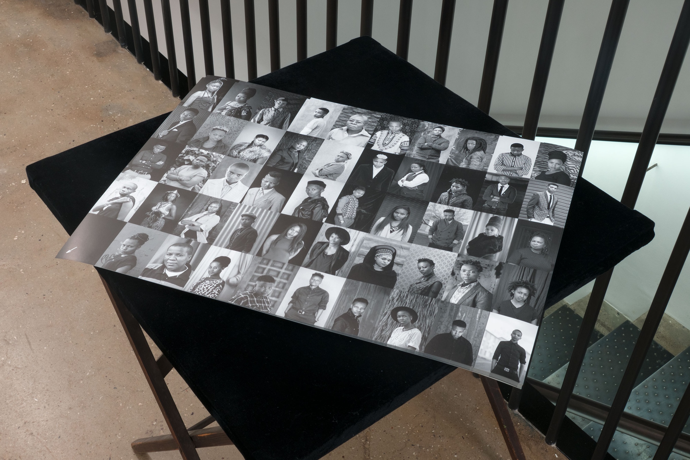 A photograph shows a monochrome litho poster with a grid of photographic portraits from Zanele Muholi's 'Faces & phases' project on a black table.
