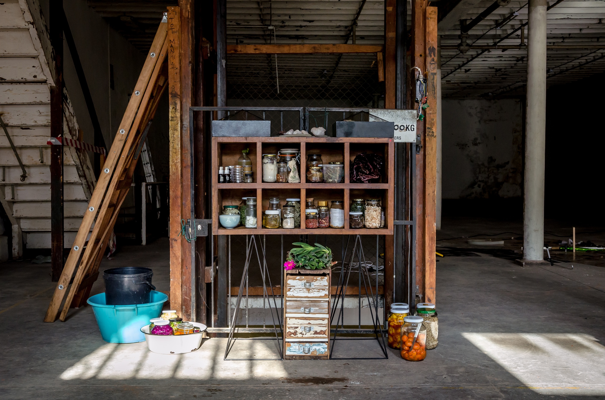 Installation photograph from ‘The Apocalypse Pantry’, Zayaan Khan and Heather Thomson’s residency on A4’s 1st floor. In the middle, a wooden shelf with bottled preserves and plant matter is mounted on a metal gate, with worn wooden drawers and more bottled plant matter sitting on the floor below it.
