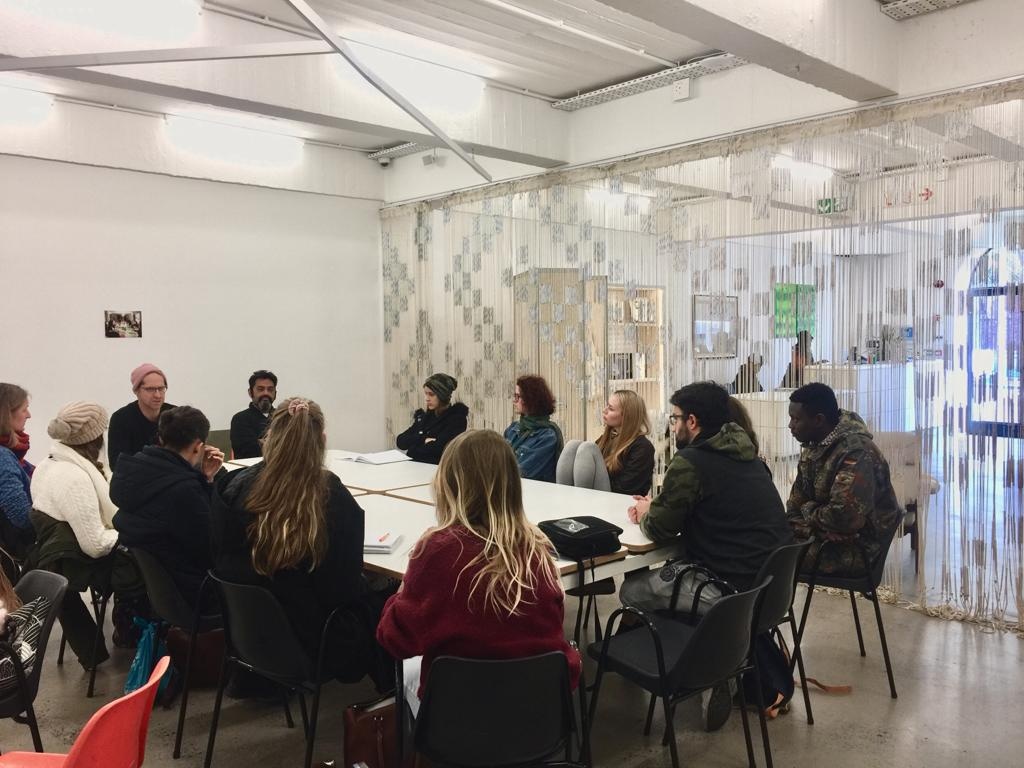 Event photograph from an exchange with Stellenbosch Academy of Design and Photography on A4’s ground floor. On the left, Josh Ginsburg and Honours students from the academy are seated around a table. On the right, Igshaan Adams’ mixed media tapestry ‘Al asma ul Husna’ is suspended from the ceiling.
