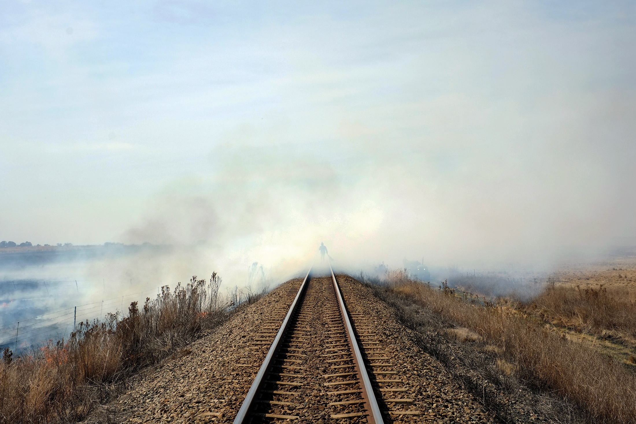 Photograph from Roger Palmer’s photo-book ‘SPOOR,’ launched on A4’s ground floor. In the middle, a railway runs off into the horizon, covered in smoke from a burning field on the lefthand side. A human figure is partially visible in the smoke.
