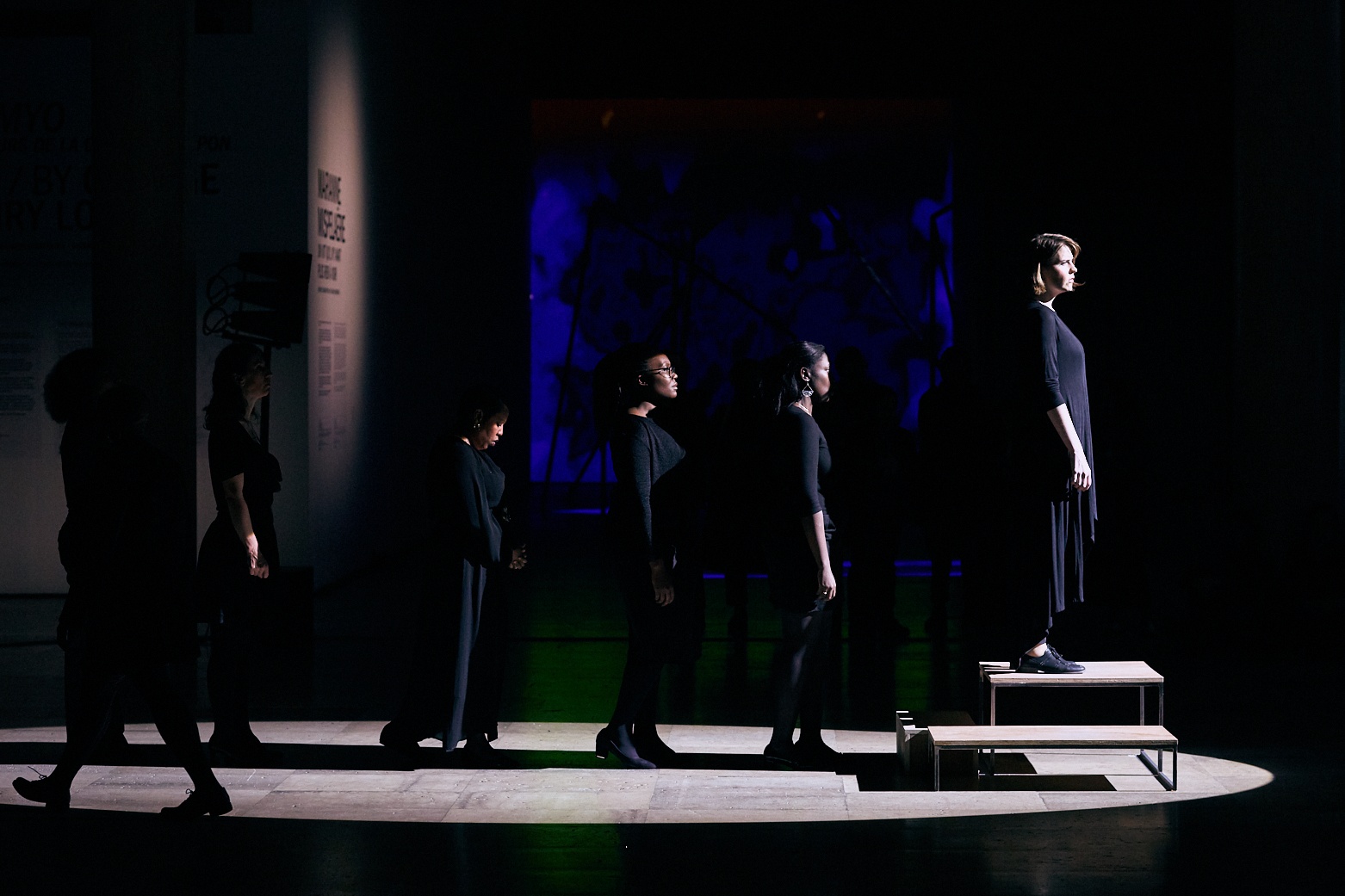 Event photograph from the offsite performance of ‘Elegy’ by Gabrielle Goliath at the D!STURB festival in Paris. A lineup of performers dressed in black stretches to a wooden podium with a performer standing on it on the right.
