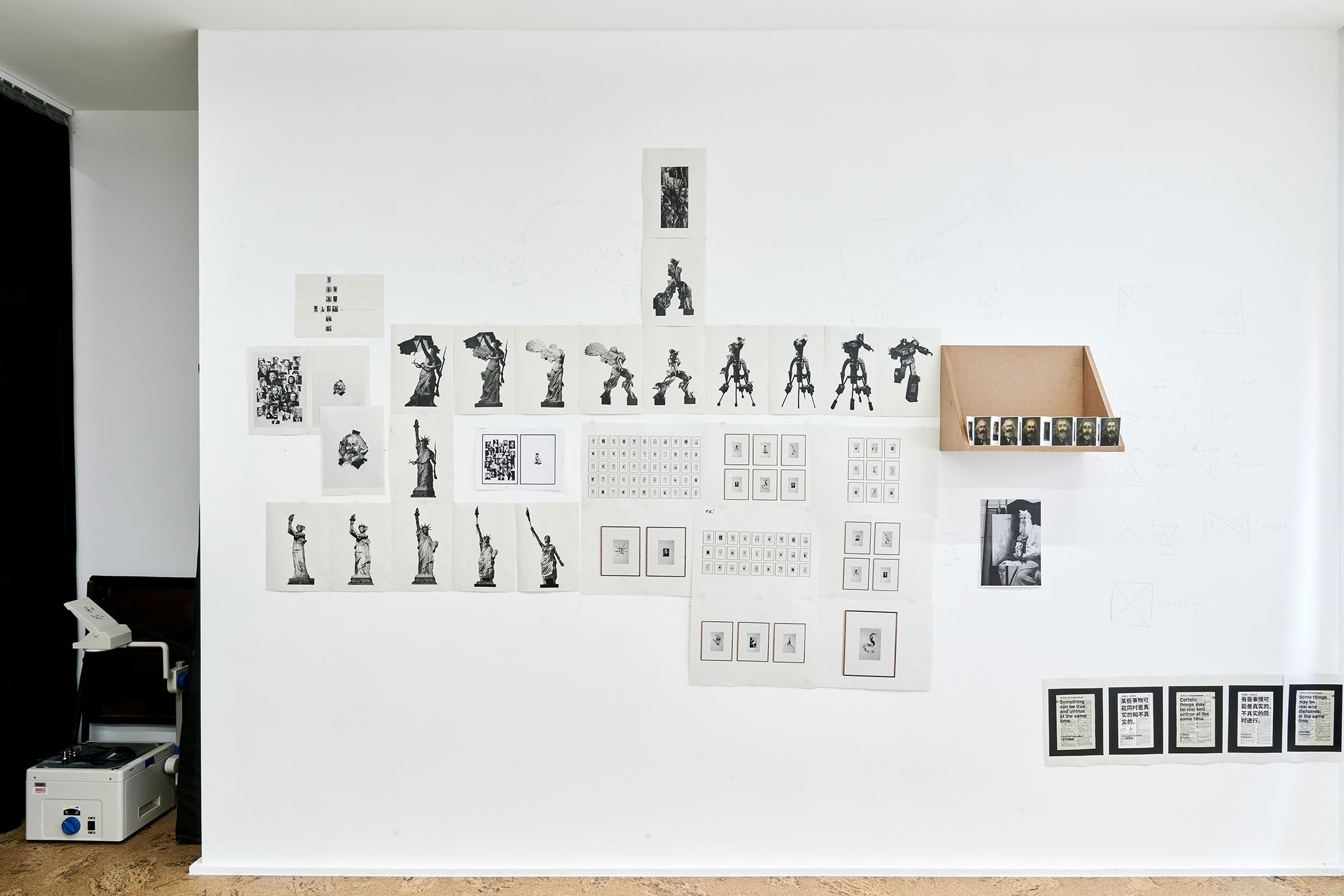 Process photograph from Patrick Waterhouse's 2022 residency in A4 Arts Foundation's studio shows photographic prints on paper arranged along the studio wall, and on a wooden wall-mounted shelf.
