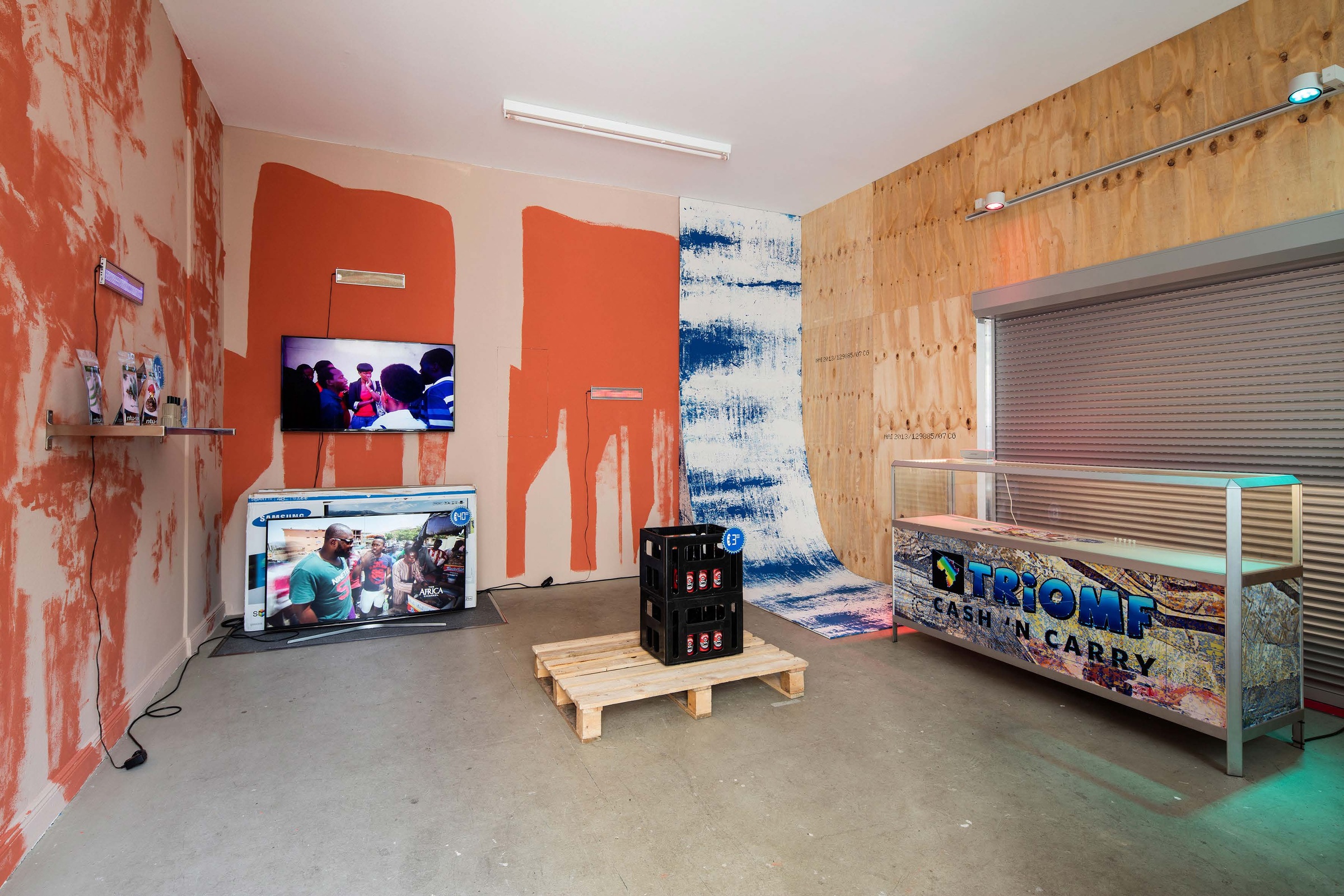 Photograph from the CUSS Group’s booth at the 2016 Berlin Biennale. On the left, printed matter is displayed on a wall mounted shelf, with the wall painted in two different shades of orange. At the back, a wall mounted screen and a screen sitting on the floor below it display video material, with the wall similarly painted. On the right, a cash ’n carry display case sits in front of a bare wood panelled wall. In the middle, a wooden pallet with a black plastic crate.
