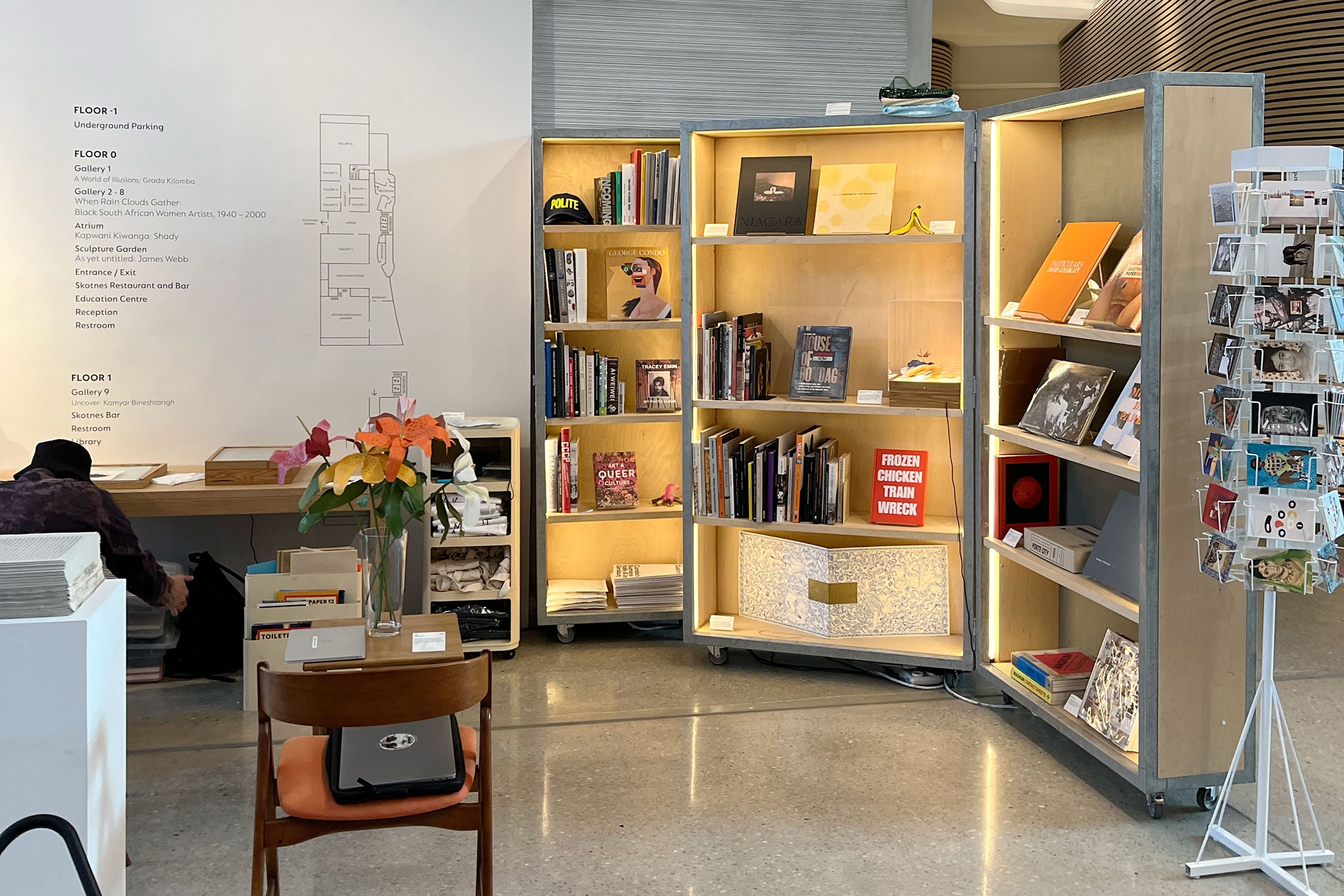 Event photograph from the Proto museum shop's booth at the 2022 Cape Town Art Book Fair. On the right, a display cabinet holds a selection of art books, alongside a revolving stand with postcards. On the left, two tables and a plinth holds art objects and publications.
