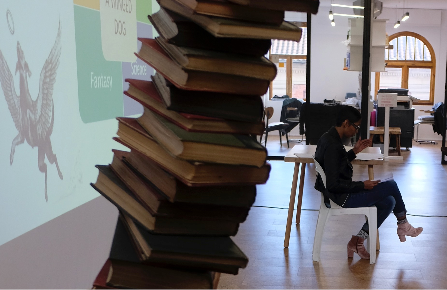 Event photograph from the Afro-futurish academic colloquium on A4’s top floor. At the back, Dr Nedine Moonsamy sits in front of a projection on the wall. At the front, a detail of Yinka Shonibare sculpture ‘Boy Balancing Knowledge II’ consisting of a stack of books leaning to the right.
