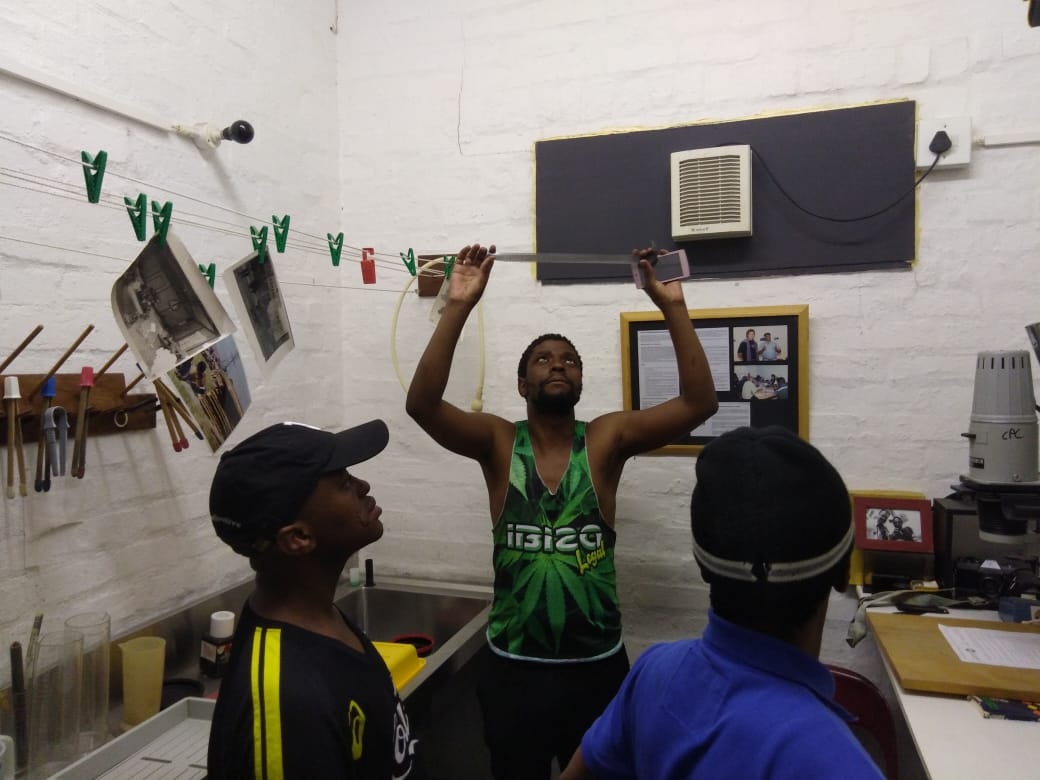 Event photograph from the ‘Open studio’ exchange on A4’s top floor that forms part of Jabulani Dhlamini and Sabelo Mlangeni’s residency. It depicts a photographic dark room, with Mlangeni standing among two other individuals, his arms raised to hold and inspect a strip of photographic negatives.
