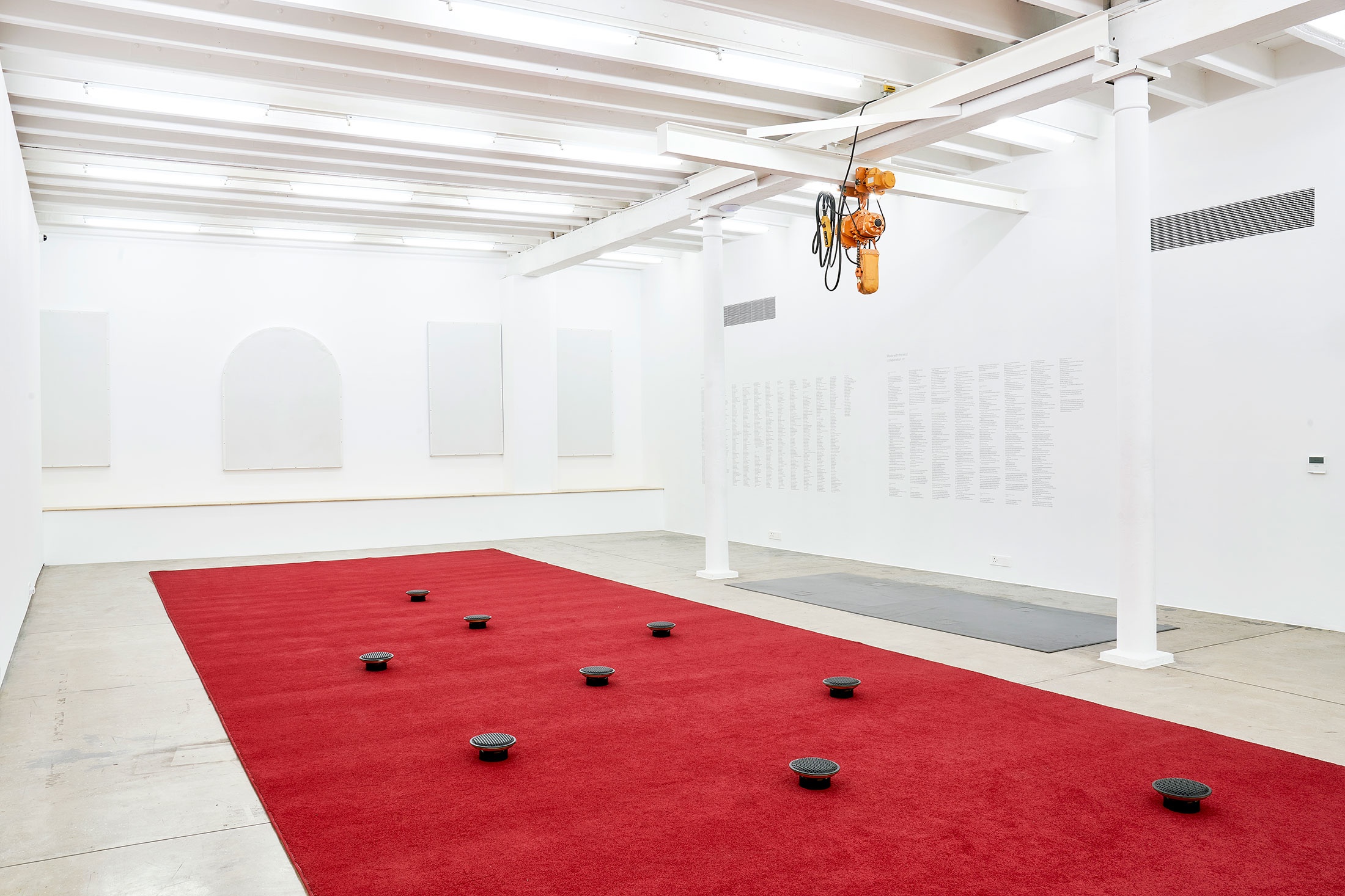 Installation photograph from The Future Is Behind Us exhibition in A4’s Gallery. In the middle, James Webb’s sound installation ‘Prayer’ features an long red carpet with upturned audio speakers laid out in a geometric pattern.
