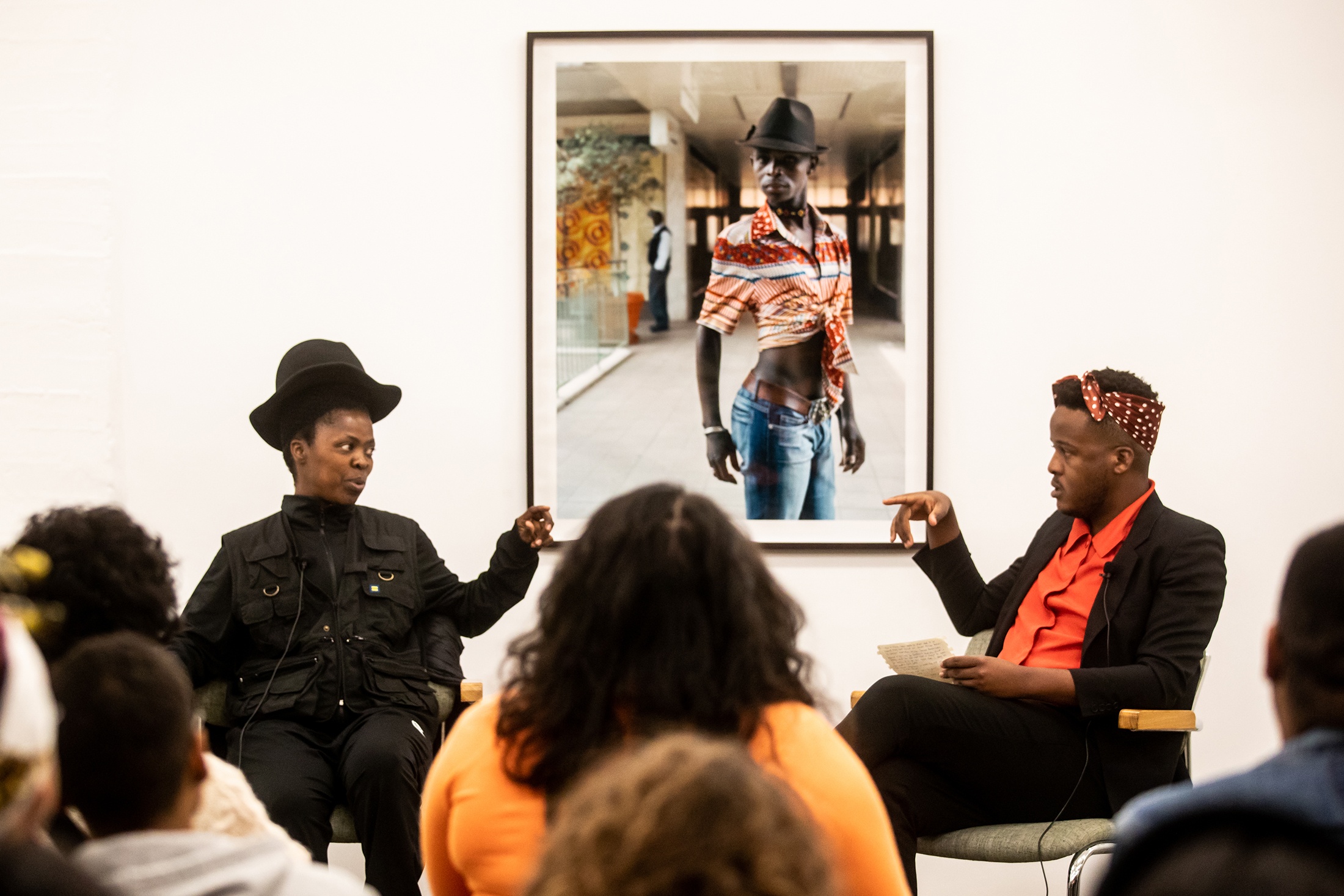 Event photograph from ‘Bare in Conversation with Sir Professor Zanele Muholi’, Session 1 of the ‘Bare Stories’ series hosted on A4’s ground floor. At the back, Muholi’s framed photograph ‘Martin Machapa’ is mounted on the white wall. In the middle, visual activist Zanele Muholi and Welcome Lishivha sit in conversation. At the front, attendees are seated.
