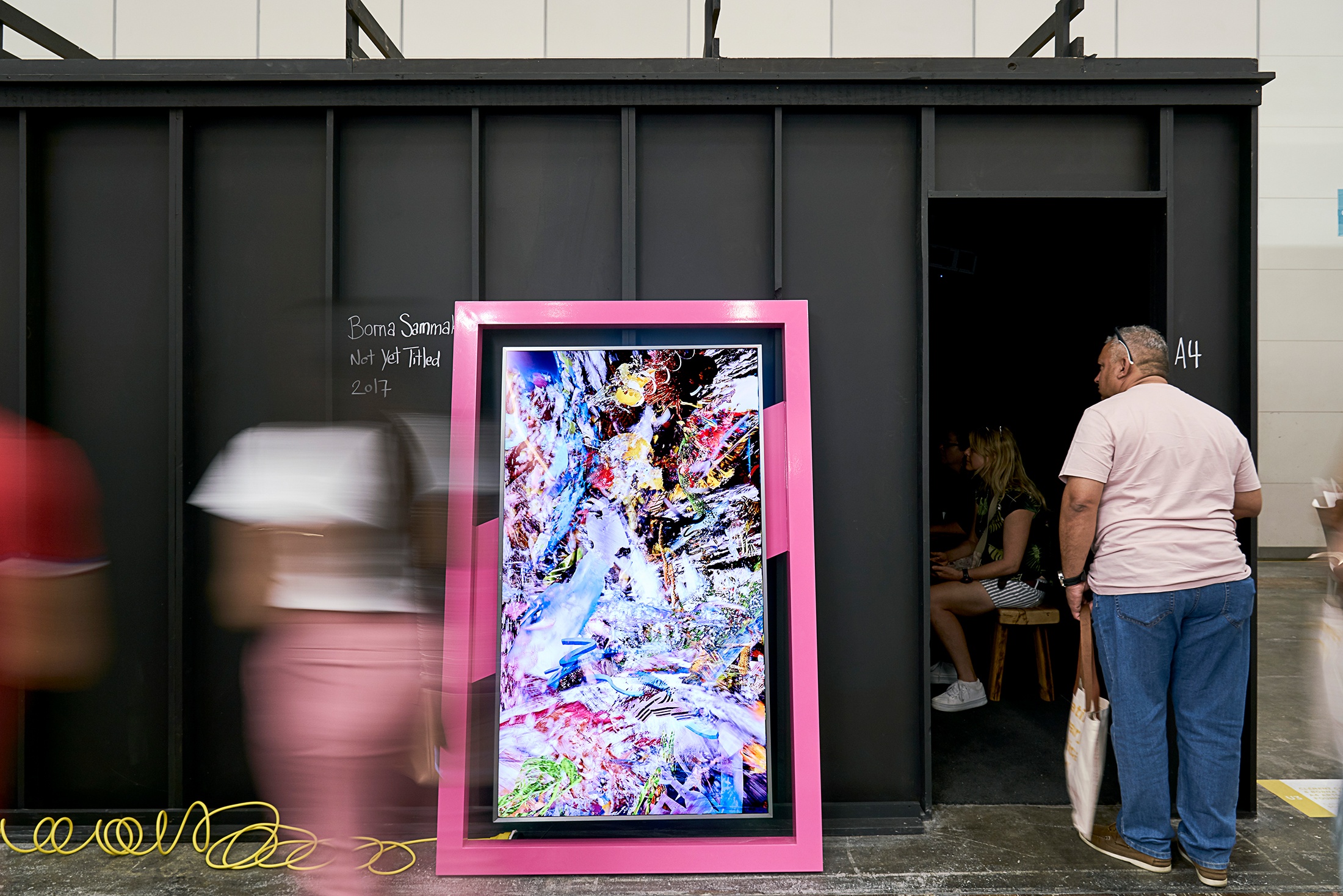 Event photograph from A4’s booth at the 2019 rendition of the Investec Cape Town Art Fair. In the middle, Borna Sammak’s video sculpture ‘Not Yet Titled’ lies against a large black metal booth.
