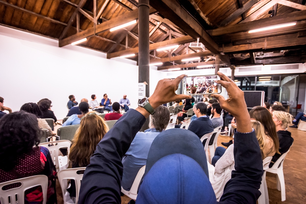 Event photograph from the book launch of Ashraf Jamal’s ‘In the World: essays on contemporary South African art’ on A4’s top floor. At the front, a seated participant holds up a phone to take a photo of the panelists. At the back, Jamal sits in conversation with Gcobani Sipoyo, Alexandra Dodd and Andrew Lamprecht.
