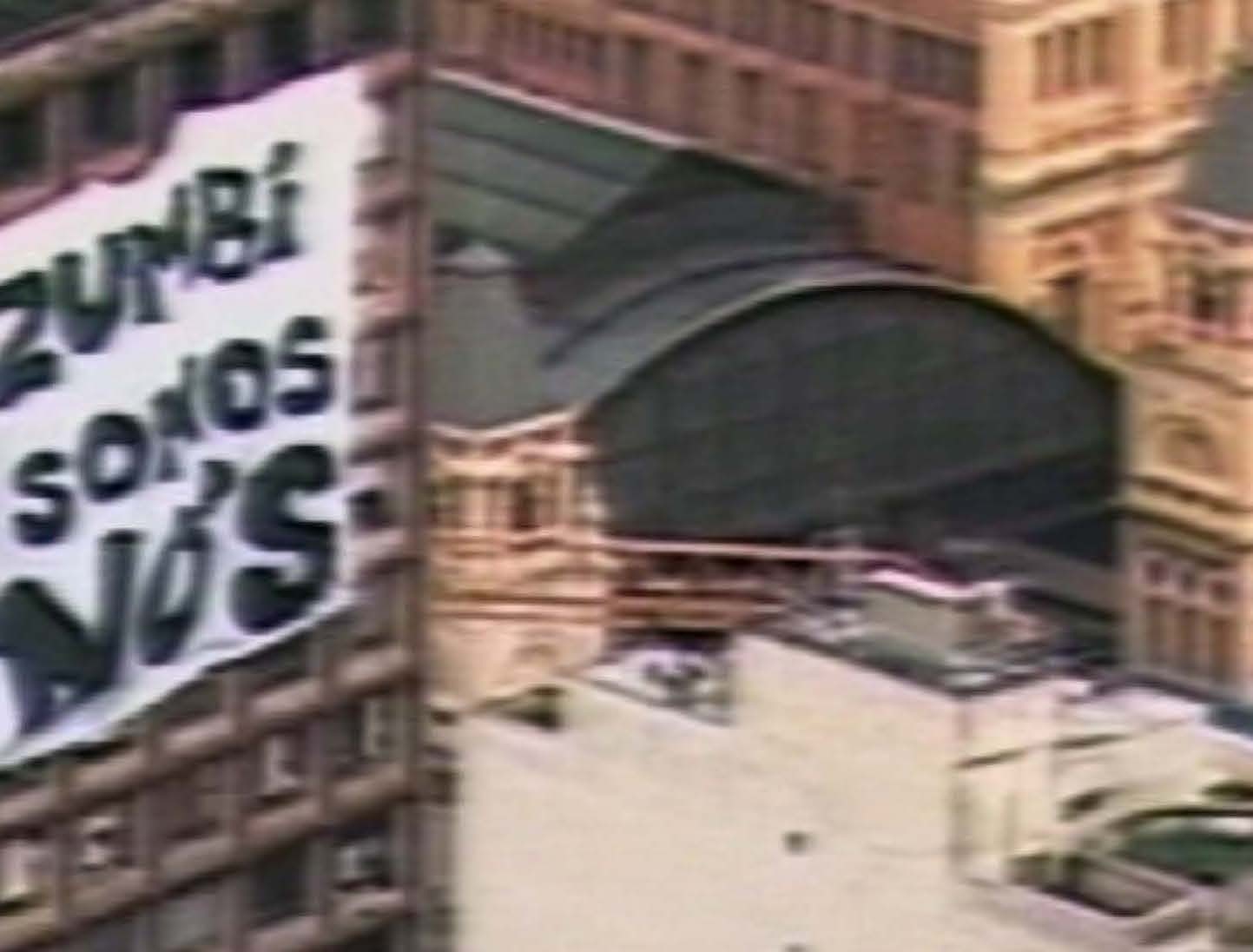 Still frame from Daniel Lima and Amílcar Patel’s documentary film ‘Zumbi Somos Nós’ (‘We are Zumbi’) screened on A4’s ground floor. An image of a cityscape with a large white banner hanging from a building on the left that reads ‘Zumbi Somos Nós’.
